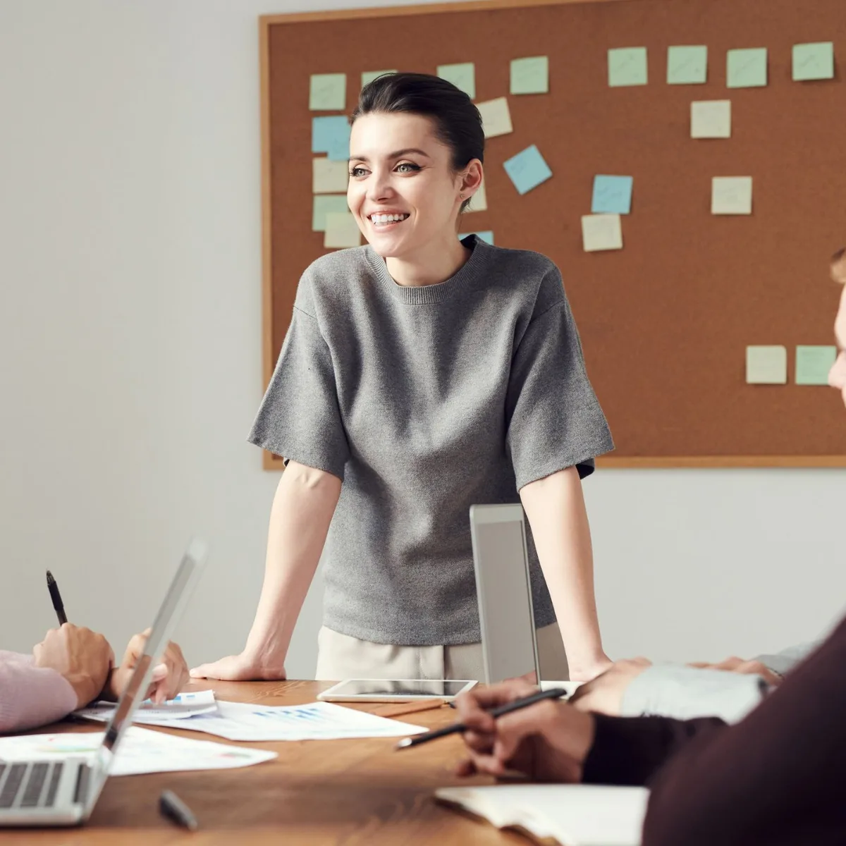 Smiling woman leading a team meeting around a table with laptops and notes, in front of a board of sticky notes
